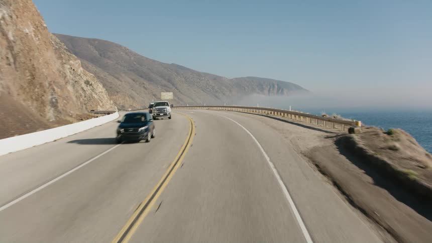 Vehicles travel an oceanside highway beside a mountain