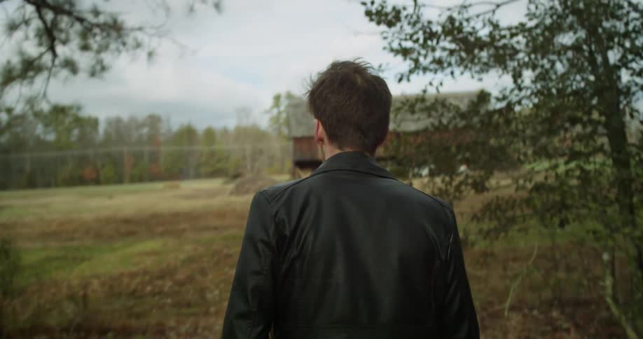 Man in a black leather jacket walks to a run down red barn,