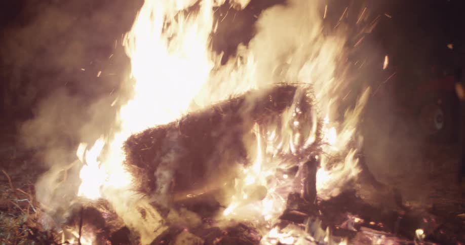 People pick up a large bale of hay and throw it in a campfire at night