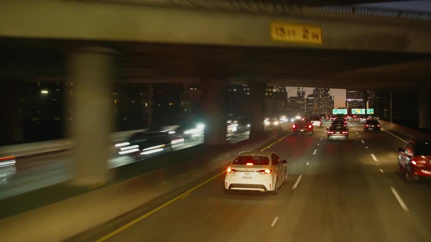 Cars drive on divided highway next to illuminated high rise buildings at dusk
