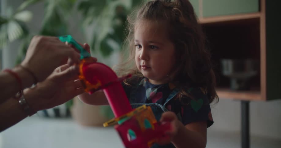 Daughter with curly hair and mother sit on floor playing with toys in living room at home