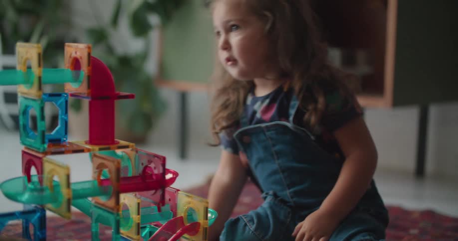 An adult hands a toy ball to a young girl who places it in a toy set and watches it roll down