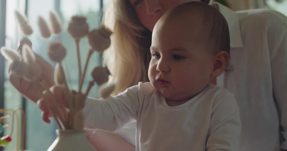 Blonde mother wearing white shirt and baby hold dry flower in living room at home