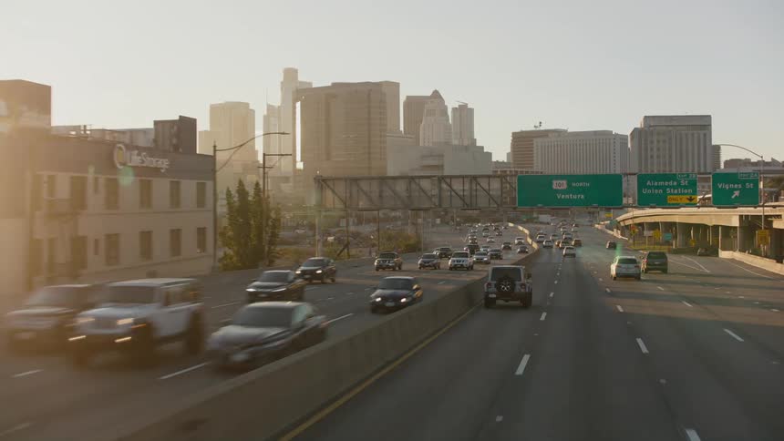 Traffic on a highway traveling downtown with the sun shining behind tall buildings and skyscrapers in the distance