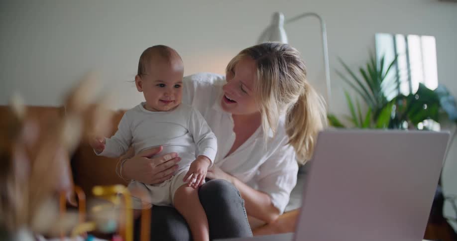 A woman rocks a baby in her lap while sitting on a sofa as the baby watches a laptop lying atop a nearby table