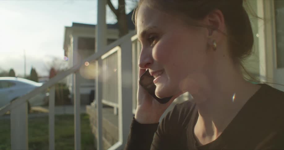 A woman is sitting outside on the front steps of a house talking on the phone