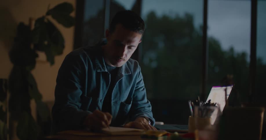Man wearing blue shirt writes in notebook and works on laptop on 3d box model in room