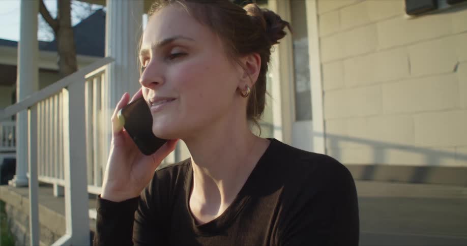 A woman is sitting outside on the front steps of a house talking on the phone
