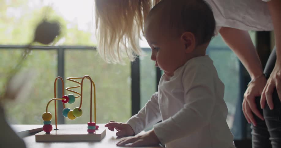 Blonde mother with white shirt and jeans holds baby playing with toys in living room at home