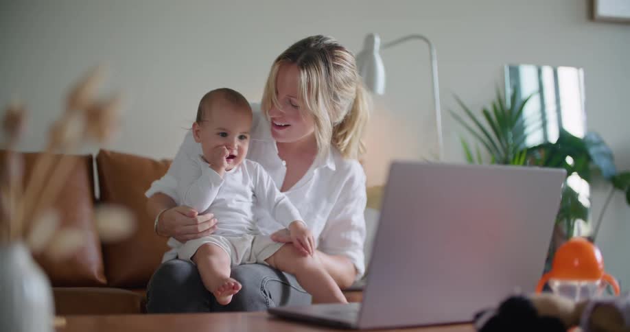 Blonde woman holds child in her lap as they sit in living room watching laptop