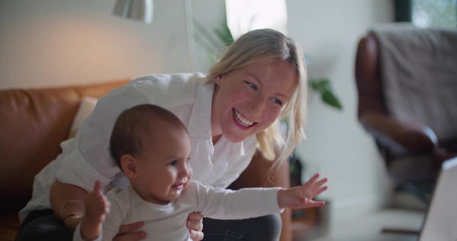 A woman attempts to get a baby to wave at a camera on a laptop while sitting indoors during the day