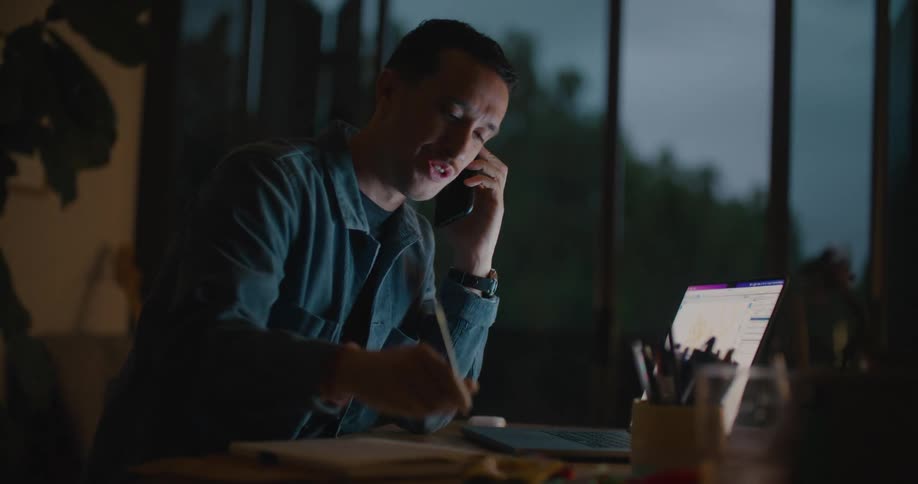 A man is sitting at a table in front of a laptop computer and talking on the phone as he writes in a notebook