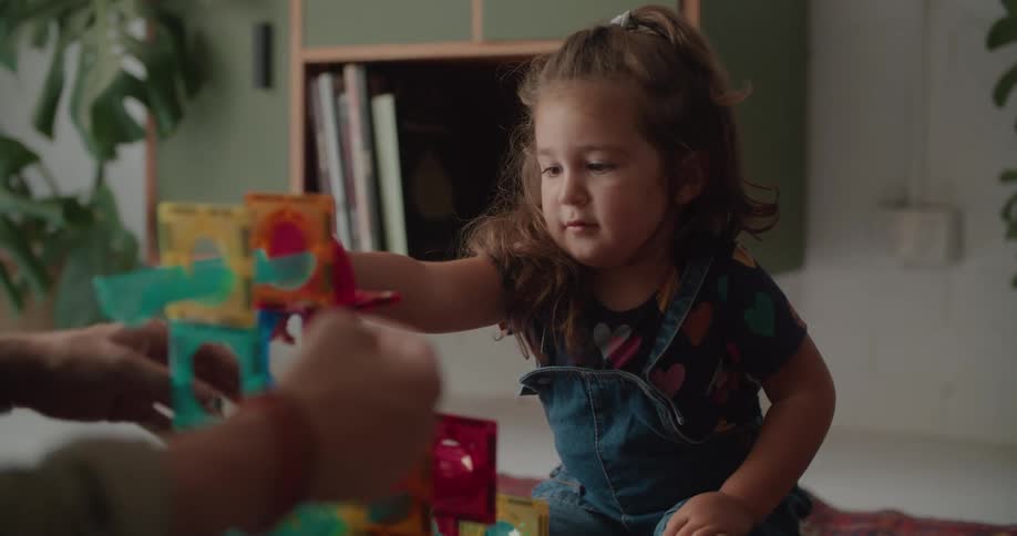 A young girl sits on the floor at home with her father and theybuild a tower out of play blocks and then they high five one another
