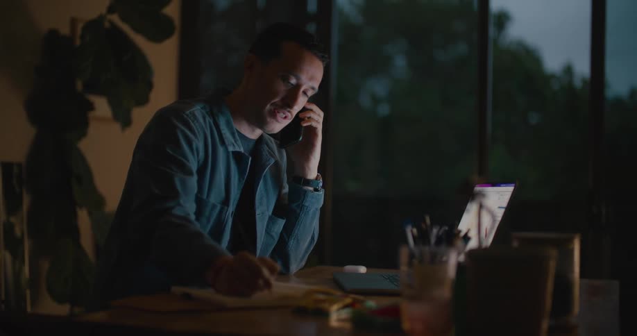 A man is sitting at a table in front of a laptop computer and talking on the phone as he writes in a notebook