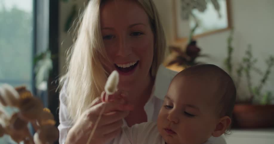 Blonde mother in white shirt holds baby playing with dry flower in living room at home