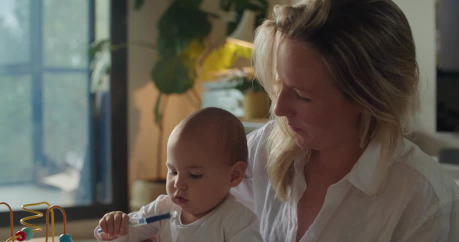 Blonde mother with white shirt holds baby drawing in notebook at coffee table in living room at home