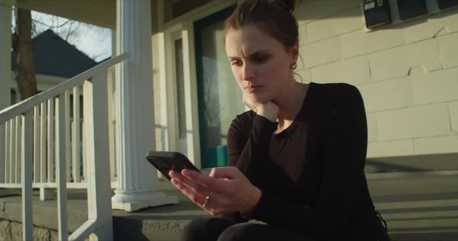 A woman sits on her front step and looks at her cell phone and then looks across the street