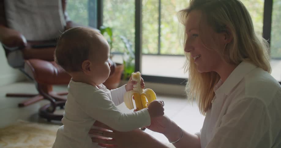 A woman at home smiles as she holds a banana and her baby takes a bite