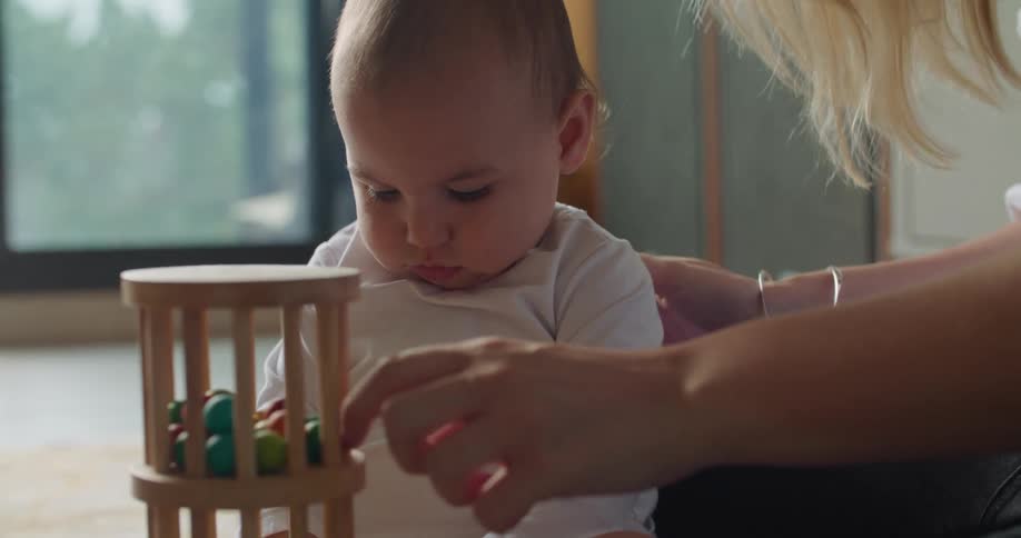 Mother turns over a wooden toy containing small colored balls as she plays with her baby on the floor