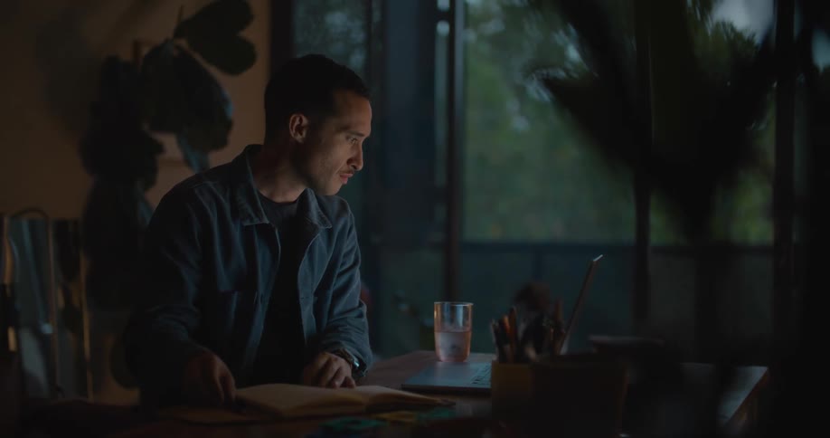 A man glances between a book and a laptop while sitting at a table indoors in the evening
