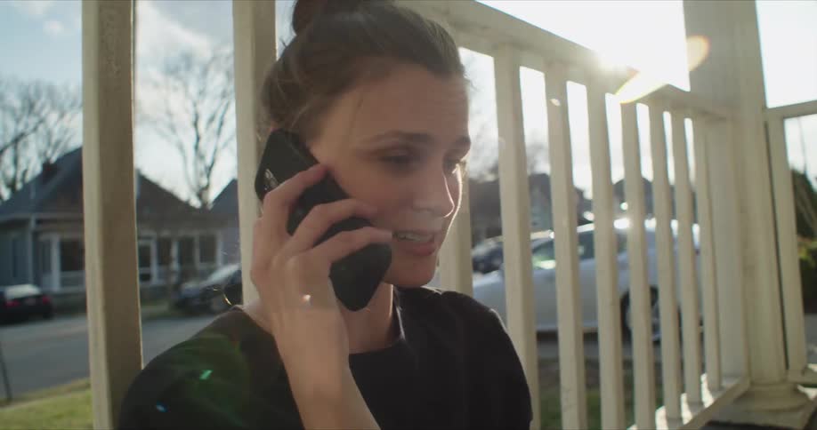 Woman dressed in black sits on a porch and talks on a cellphone as cars sit parked across the street