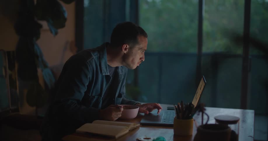 Man sits at a table and eats food from a bowl as he looks at a laptop