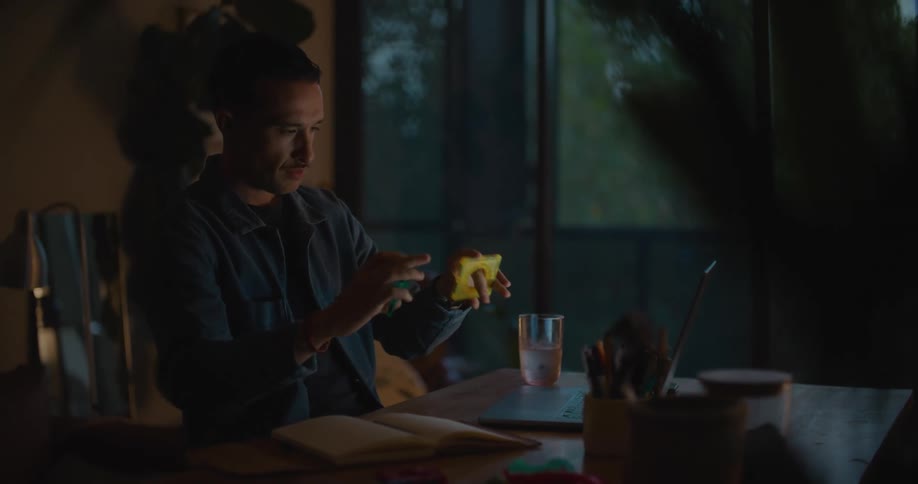 A man is sitting at a table with a laptop computer and looking at a square objects and the computer screen and writing in a book
