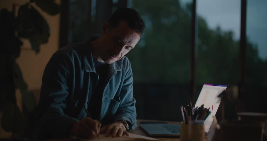 Man sitting at the desk using his laptop and writing down on a notebook