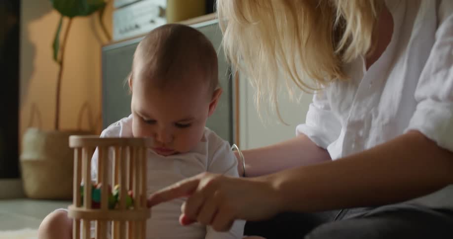 Blonde mother with white shirt and bracelets and son sit on floor playing with toys in living room at home