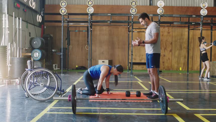 A woman exercises on a mat in a gym while being instructed by a trainer standing near her
