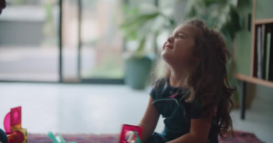 A man is sitting on the floor with a little girl holding a toy and he bends down and shakes his head near her face and she smiles and covers her eyes