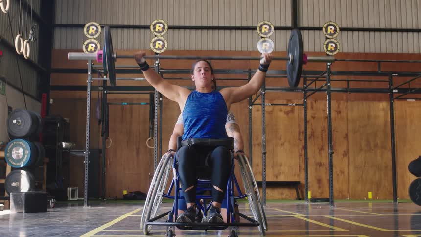 Woman in wheelchair at a gymnasium performing multiple repetitions of the overhead press exercise with a heavy barbell