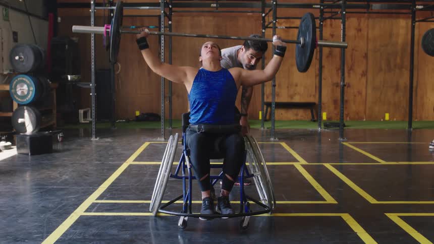 A woman in a wheelchair lifts weights in a gym while a spotter stands behind her