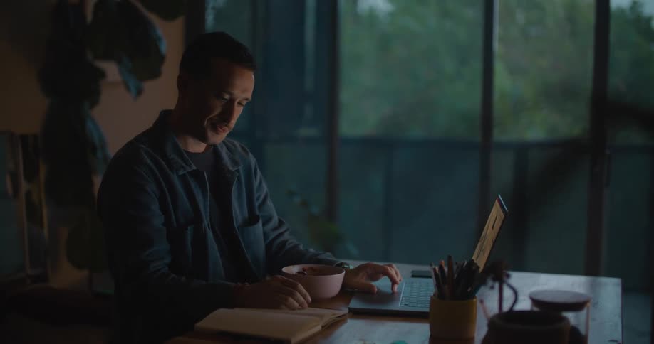 Man sits at a table and smiles as he looks at a book then types on a laptop