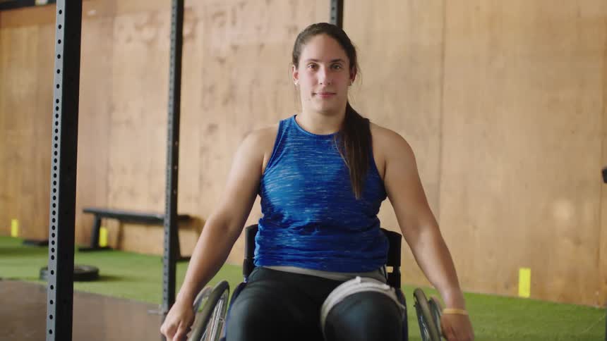 Woman wheels her wheelchair forward to sit among metal poles in gym with a smile