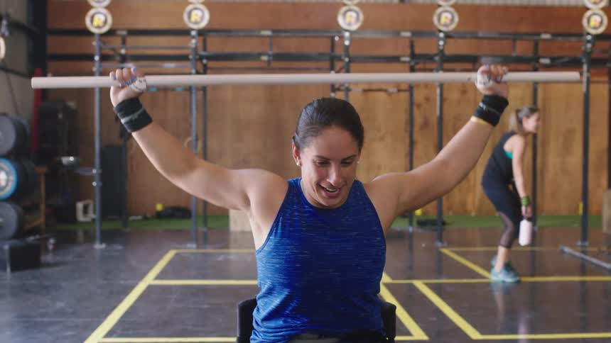 Woman in a blue tanktop raises a long white bar behind her head while in a gym