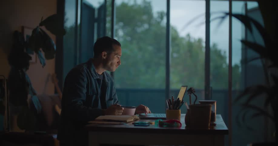 A man is sitting in at a table near a window looking at his laptop computer as he eats