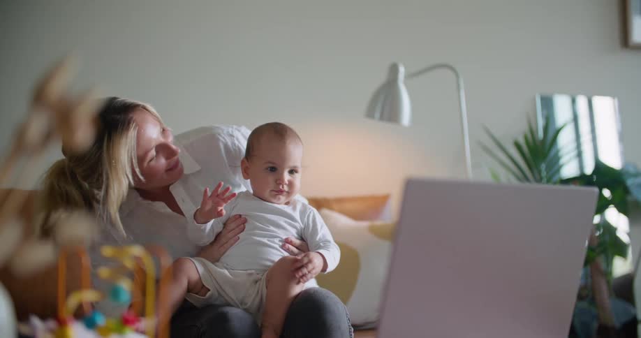 Blonde mother with shirt and pants holds baby sitting on brown couch waving at person on laptop in living room at home