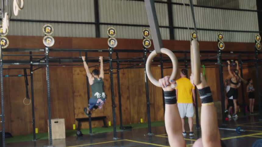 A woman exercising pulls herself up on rings as people exercise behind her