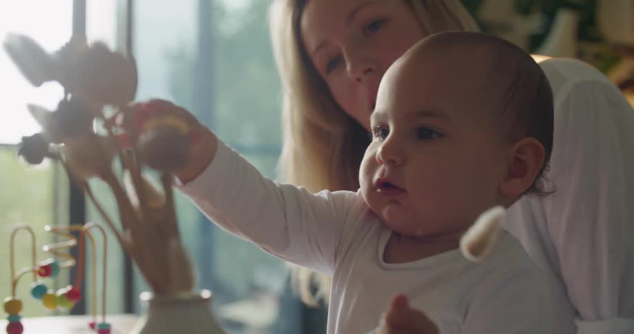 Blonde mother with white shirt holds baby playing with dry flower in living room at home