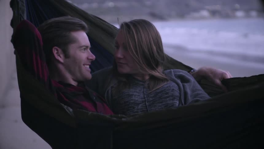 A man and woman lay in a hammock together by the water