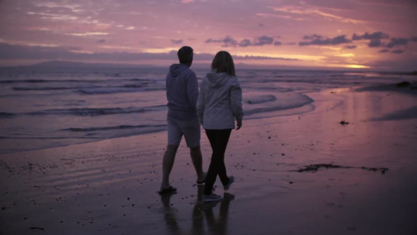 A couple walks down a beach at sunset