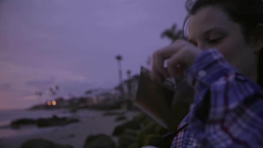A woman pulls a polaroid picture from her camera on the beach at sunset