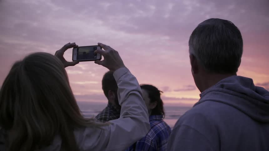 A woman takes a photo of a couple with her phone on the beach at sunset