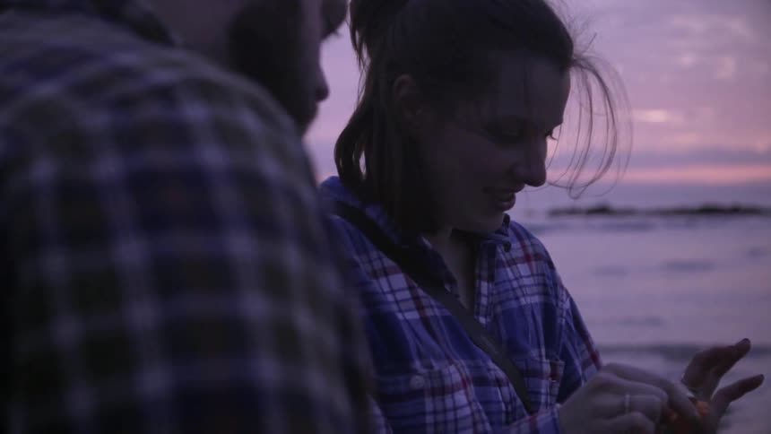 A couple standing along the shoreline as the woman brushes sand from a shell and the man watches