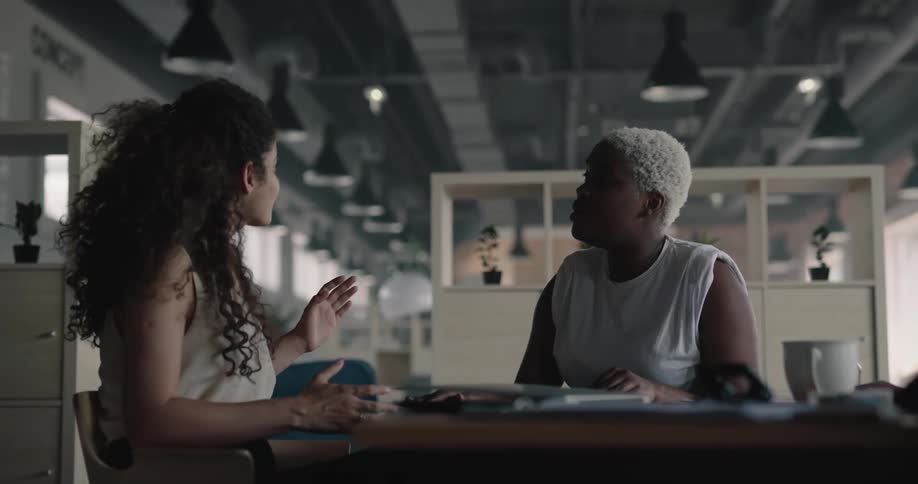 Two female coworkers sitting at the table on an office space having a conversation and laughing together