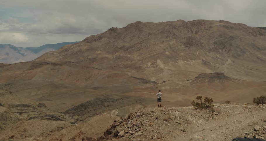 A jeep is parked on a desert mountain road and a man is standing on the edge of a cliff overlooking a valley and mountains in the distance