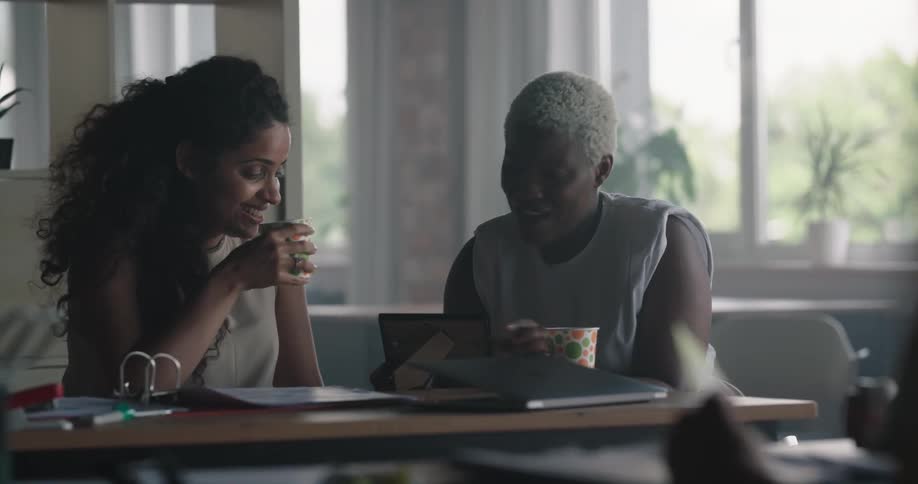 A pair of women laugh while looking at a photograph while drinking together at a table indoors