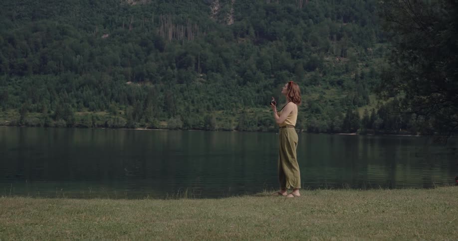 Woman with wavy brown hair yellow shirt and green pants walks on lake bank and photographs water and mountain with forest
