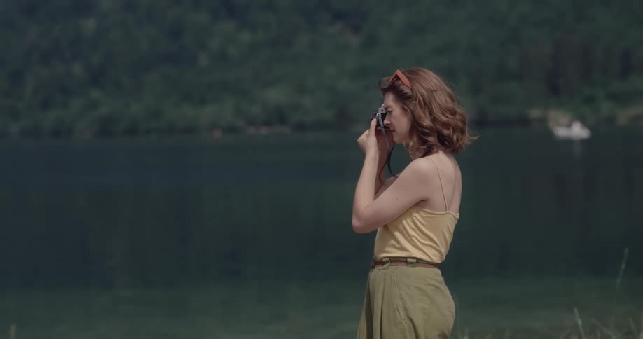 Red haired female photographer standing on shore takes pictures with boat floating near opposite forest covered shore